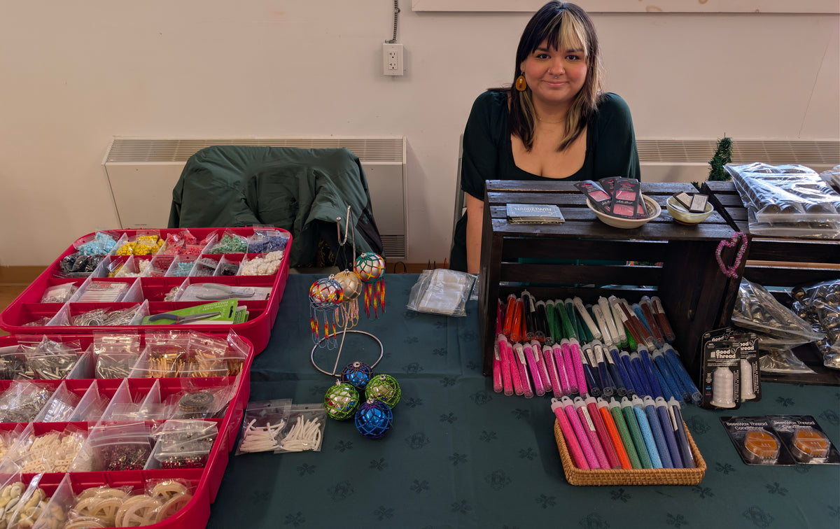 A woman with dark hair sits behind a table displaying a variety of colorful handmade crafts, beads, paintbrushes, and small packaged items in a workshop setting.
